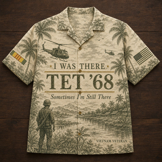 A short-sleeve button-up Hawaiian-style shirt laid flat on a dark wood background. The shirt has a vintage military-themed print in muted green-gray tones, featuring palm trees, helicopters, a soldier, an American flag, and the text 'I WAS THERE TET'68', 'Sometimes I'm Still There', and 'VIETNAM VETERAN'.